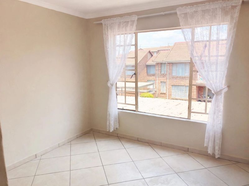 A bright, empty bedroom featuring neutral tiled flooring and a large window with views of the surrounding townhouse complex.