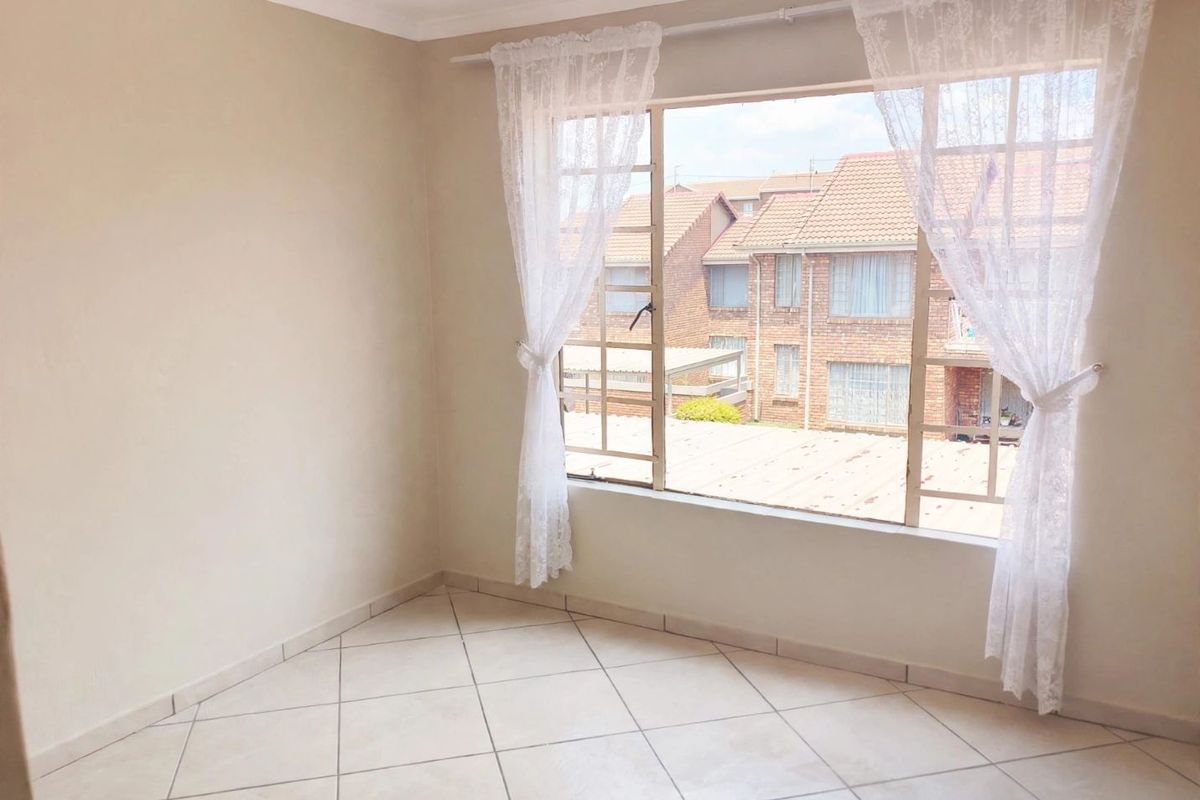 A bright, empty bedroom featuring neutral tiled flooring and a large window with views of the surrounding townhouse complex.