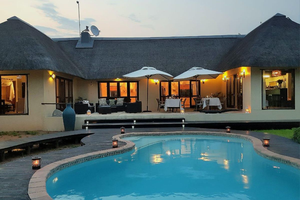 A well-maintained thatched-roof property featuring a spacious patio area overlooking a swimming pool with integrated decking and exterior lighting.