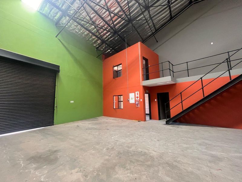 Spacious industrial warehouse unit featuring a high-clearance roof, roller shutter door, concrete flooring, and a built-in mezzanine office structure with staircase access.