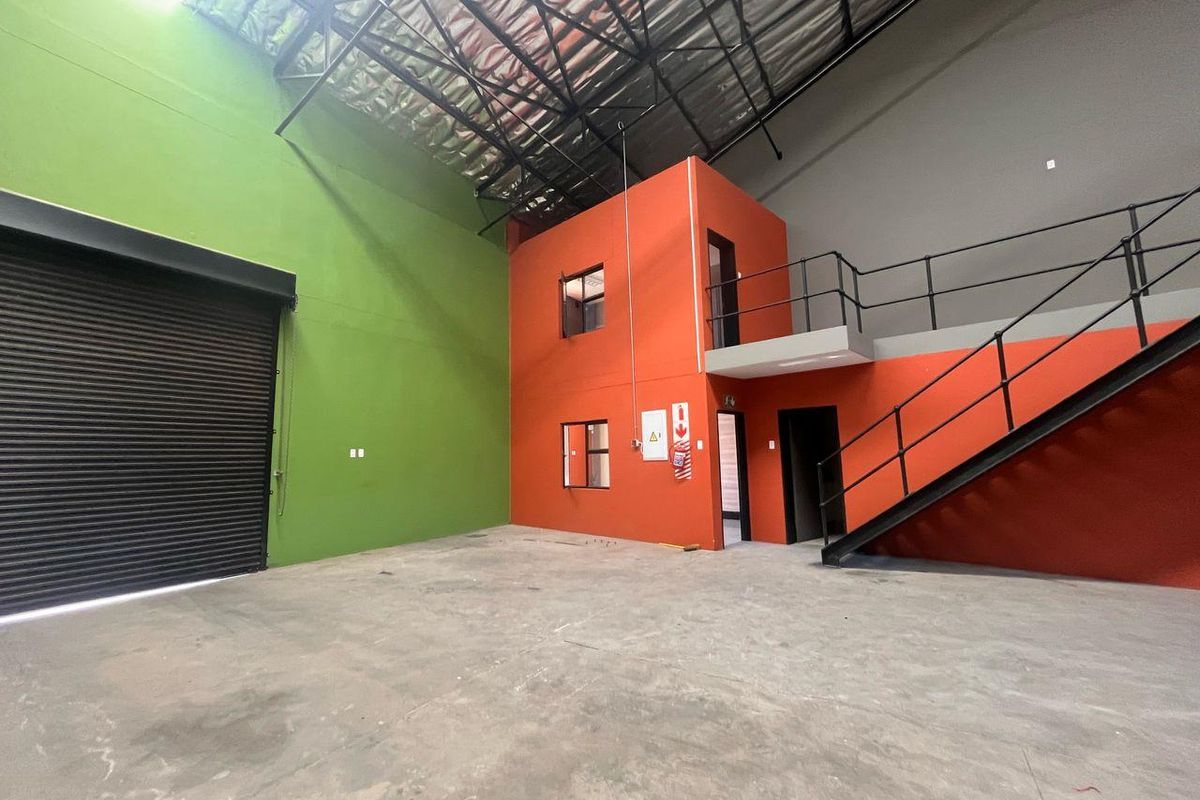 Spacious industrial warehouse unit featuring a high-clearance roof, roller shutter door, concrete flooring, and a built-in mezzanine office structure with staircase access.