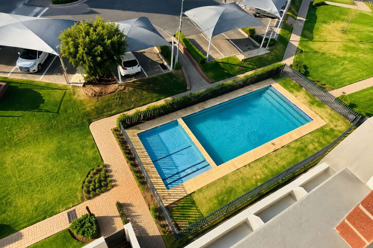 Aerial view of the apartment complex featuring a well-maintained swimming pool, landscaped garden areas, and shaded parking bays.