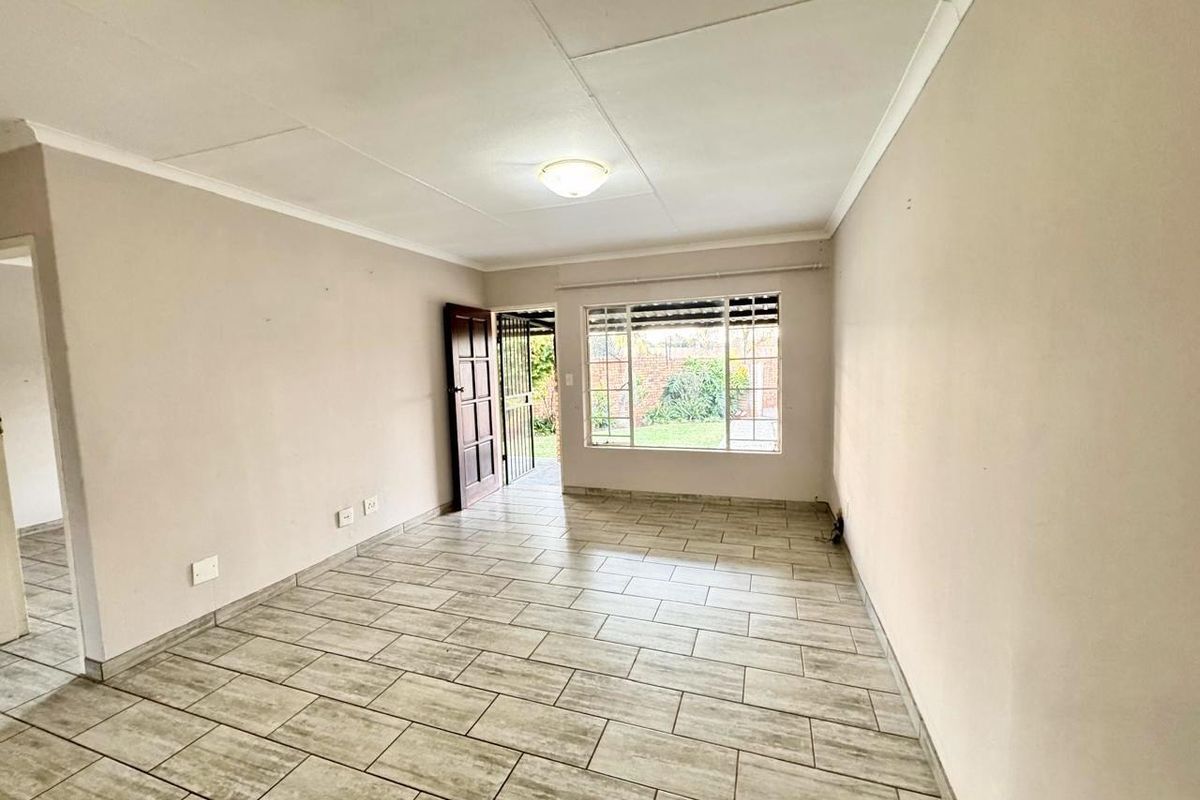 A bright, empty living room featuring neutral tiled flooring, a large window with garden views, and direct access to the exterior via a security gate.
