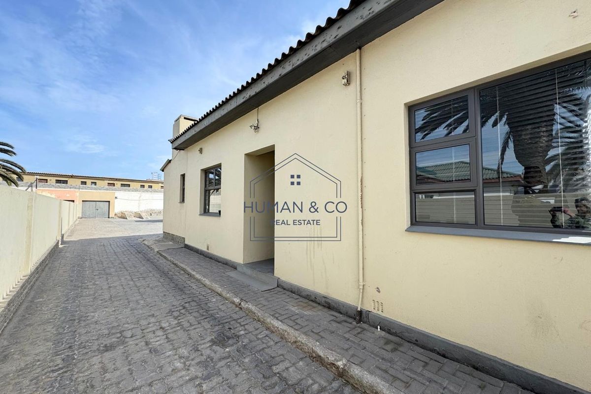 Exterior view of the property showing a paved driveway, boundary wall, and building facade with aluminium window frames.