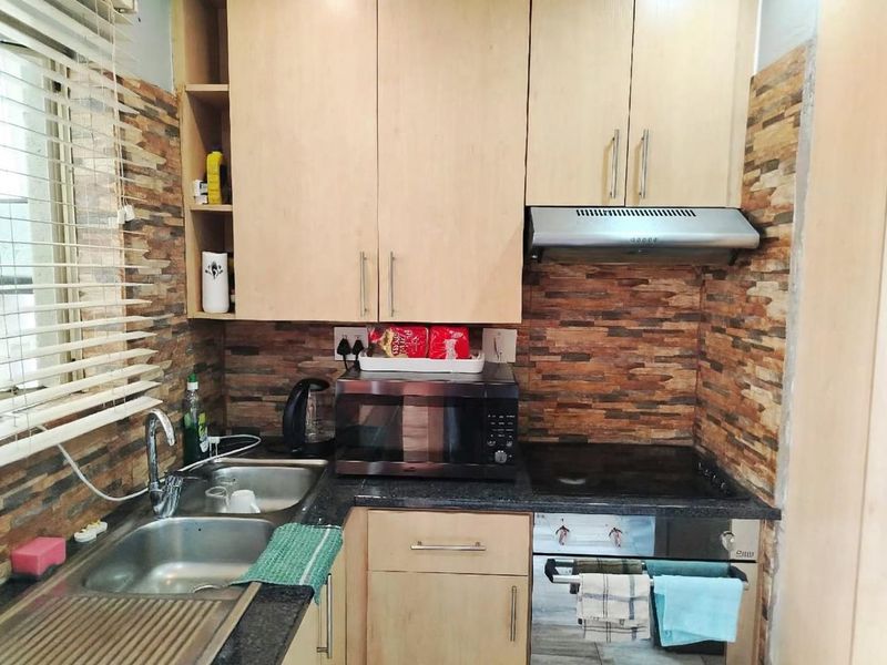 Compact kitchen featuring light wood cabinetry, stone-look wall cladding, integrated oven and hob, and a stainless steel sink with a window view.