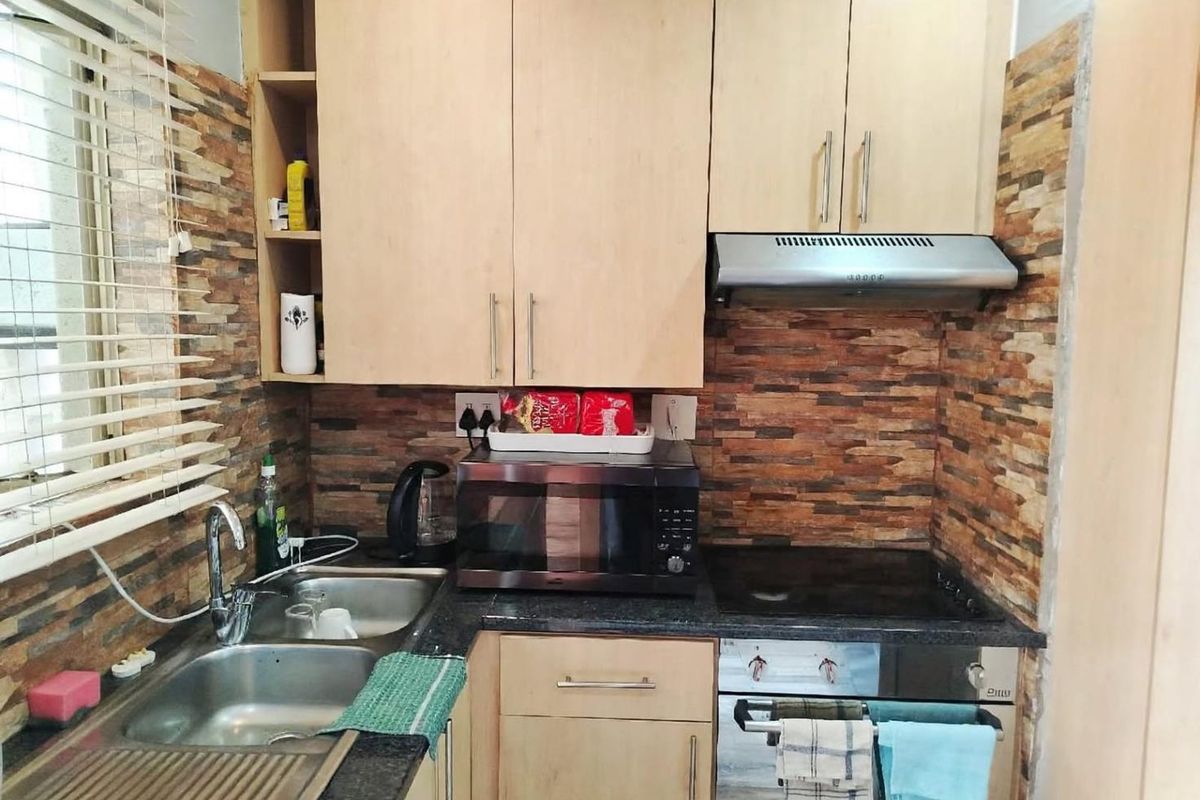 Compact kitchen featuring light wood cabinetry, stone-look wall cladding, integrated oven and hob, and a stainless steel sink with a window view.