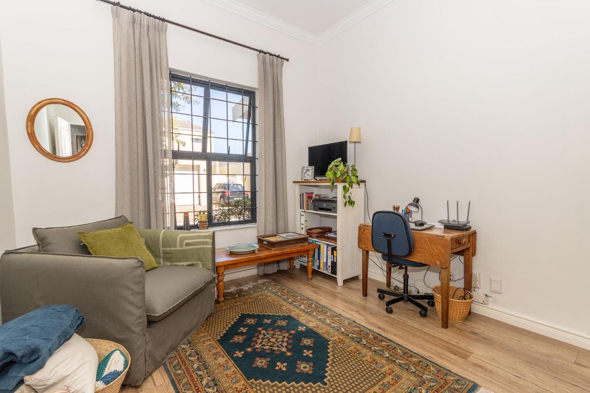 A functional bedroom or home office space featuring laminate flooring, neutral walls, and a window fitted with security bars.