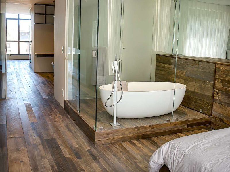 Modern bedroom suite featuring a luxurious freestanding bathtub, glass-enclosed shower, and rich timber flooring with floor-to-ceiling windows.