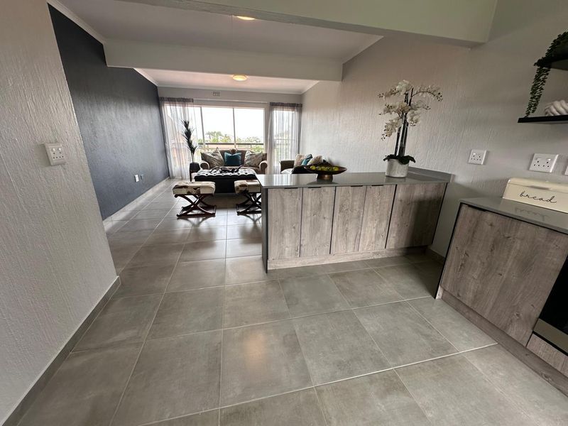Modern open-plan living area featuring large format floor tiles, a central kitchen island with wood-look cabinetry, and sliding doors leading to a balcony.