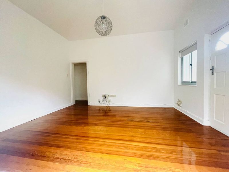 A bright, empty living room featuring polished hardwood timber flooring, neutral white walls, and a classic panelled entrance door.