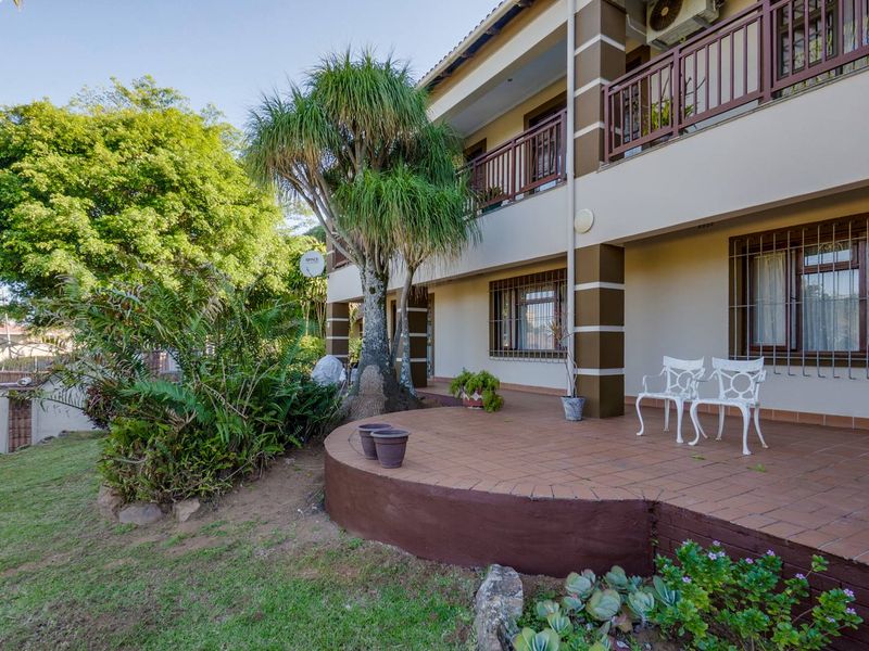A two-story residential exterior featuring a tiled ground-level patio, upper-level balcony with railings, and established garden landscaping.