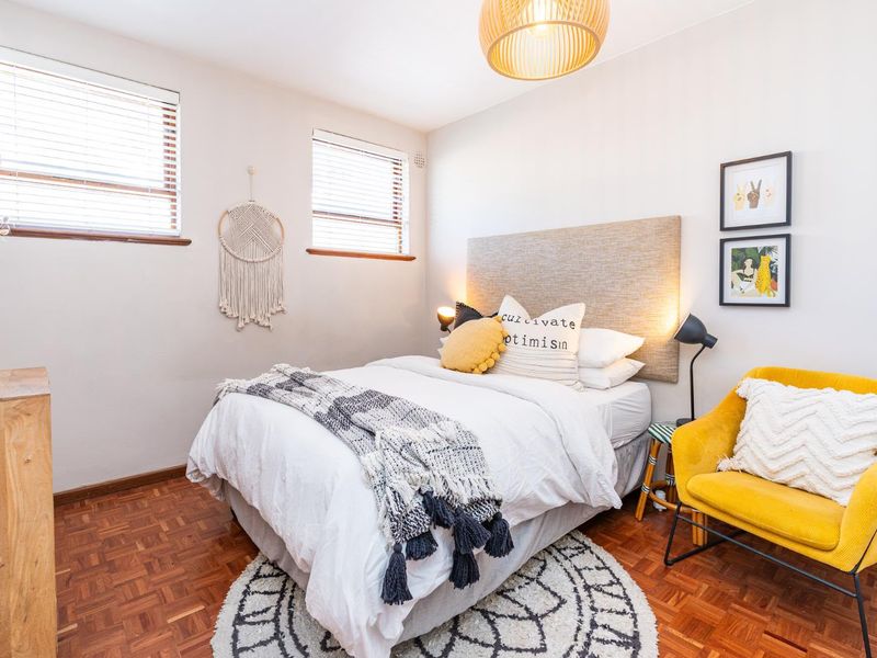 A well-lit bedroom featuring classic parquet flooring, neutral walls, and dual windows fitted with venetian blinds.
