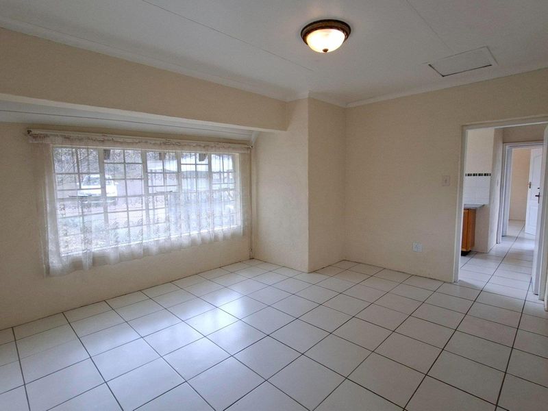 A bright, empty living room featuring neutral tiled flooring, a coved ceiling, and a large window with security bars, leading into an adjacent kitchen area.