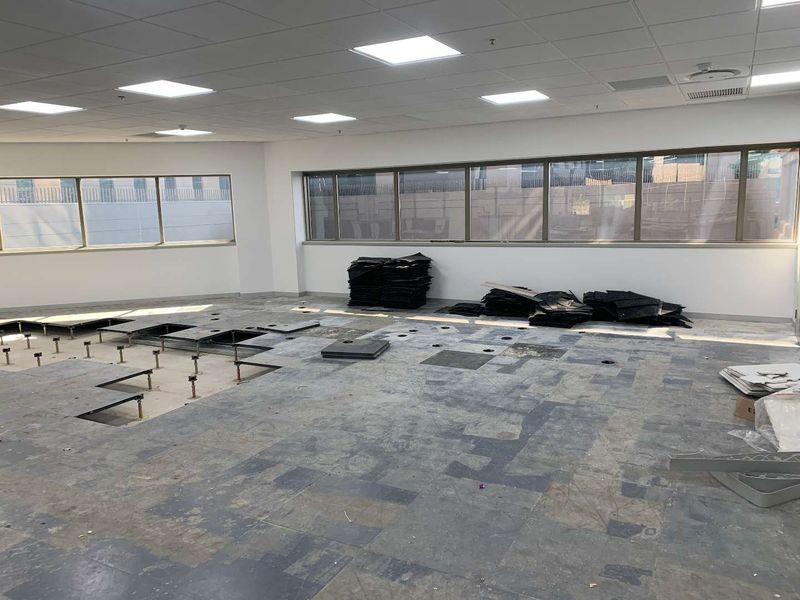 An open-plan office space currently undergoing renovations, featuring a suspended ceiling, recessed lighting, and an incomplete raised access flooring system.
