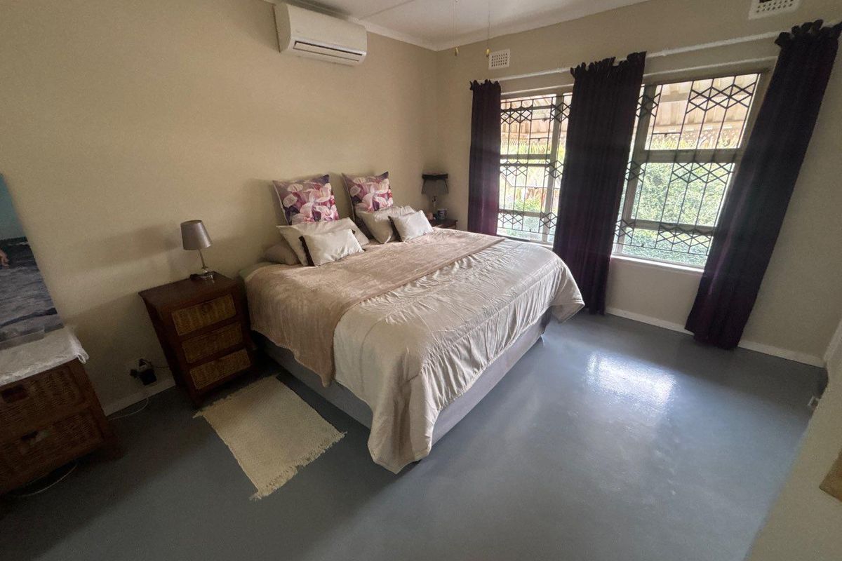 A spacious bedroom featuring painted concrete flooring, a wall-mounted air conditioning unit, and large windows fitted with security bars.