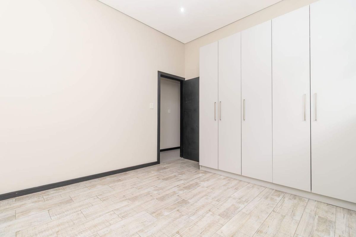 A bright, empty bedroom featuring modern white built-in cupboards, light wood-look tiled flooring, and a clean, neutral aesthetic.