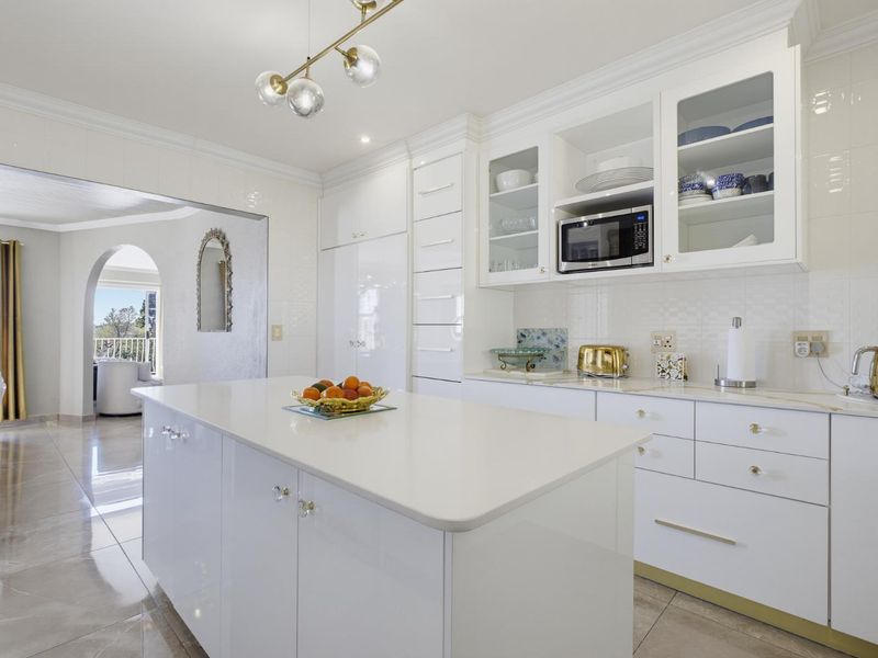 Modern white kitchen featuring a central island, quartz countertops, built-in microwave, glass-front cabinetry, and elegant pendant lighting, seamlessly flowing into the adjacent living area.