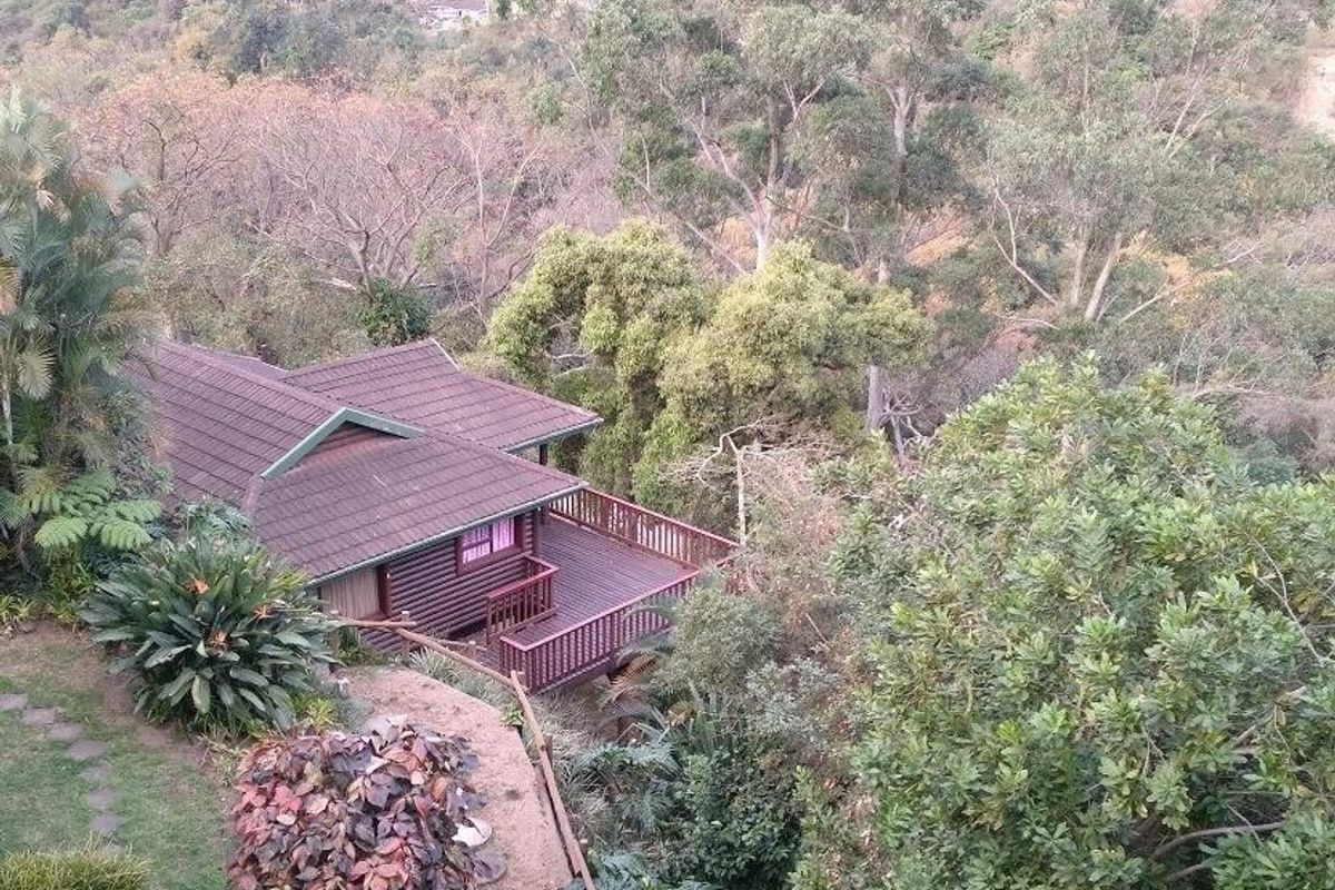 An elevated aerial view of a house featuring a tiled roof and a wooden deck, nestled within a lush, tree-filled landscape.