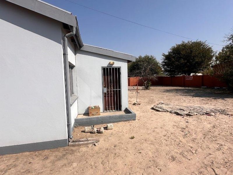 Rear exterior view of the house showing a side entrance with a security gate, downpipe, and a large, undeveloped sandy backyard area.