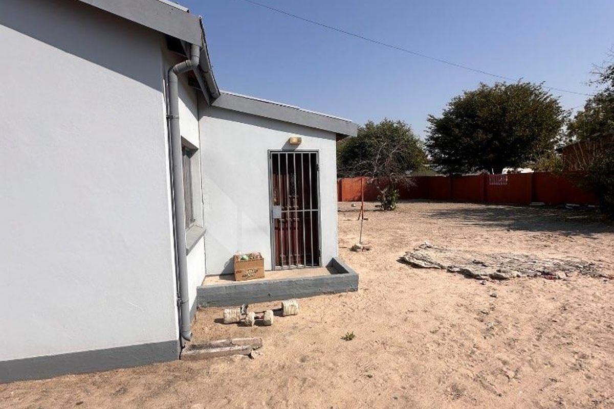 Rear exterior view of the house showing a side entrance with a security gate, downpipe, and a large, undeveloped sandy backyard area.