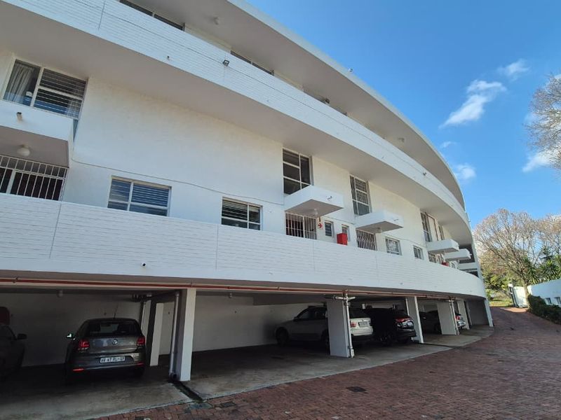 Exterior view of a multi-story apartment building featuring covered ground-level parking, private balconies with security bars, and paved driveway access.