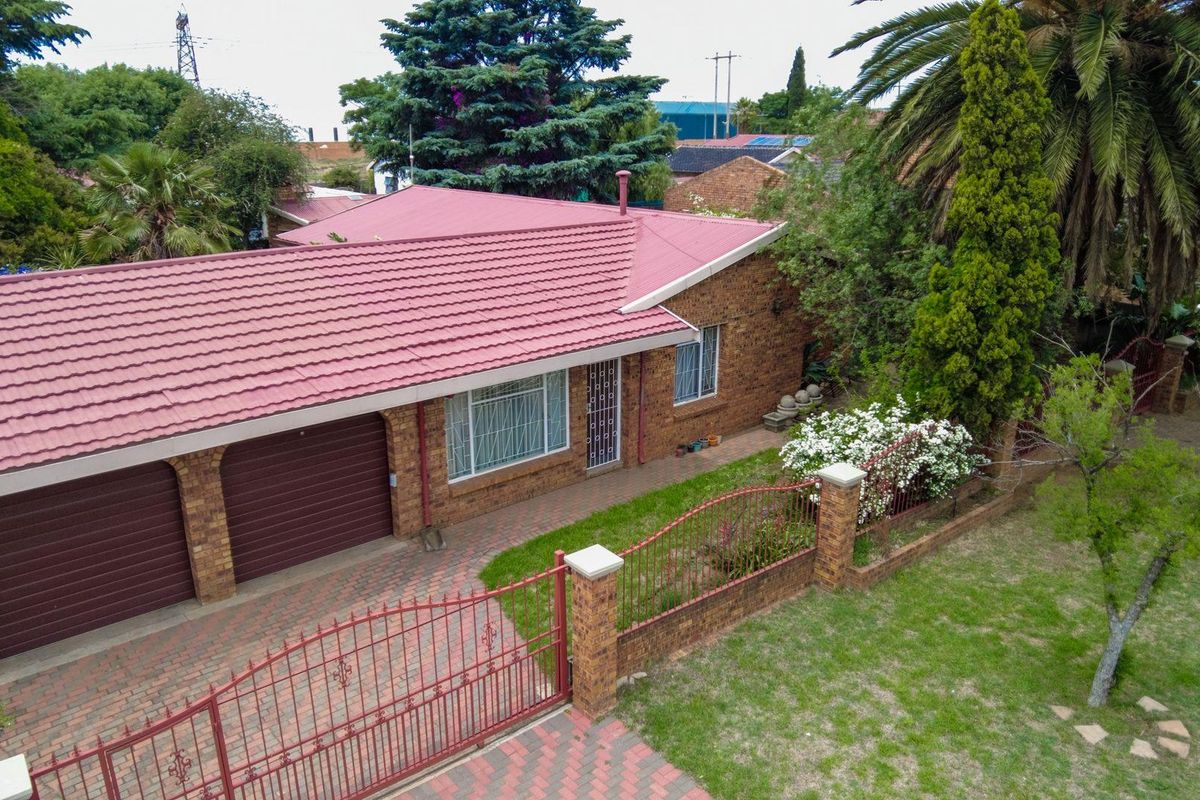 Aerial view of a brick-faced house featuring a red metal roof, double garage, paved driveway, and a gated front garden.