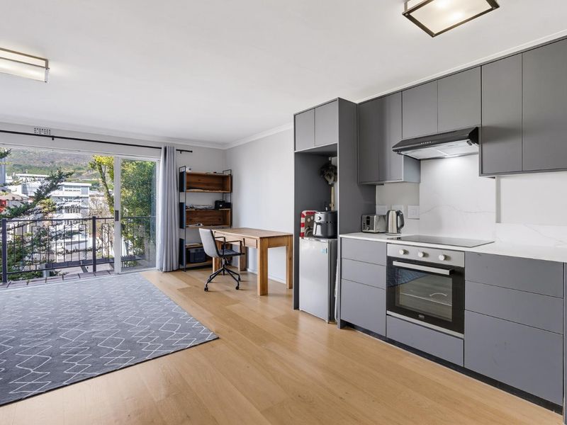 Modern open-plan apartment featuring a sleek grey kitchen with integrated appliances, light wood flooring, and a sliding door leading to a private balcony.