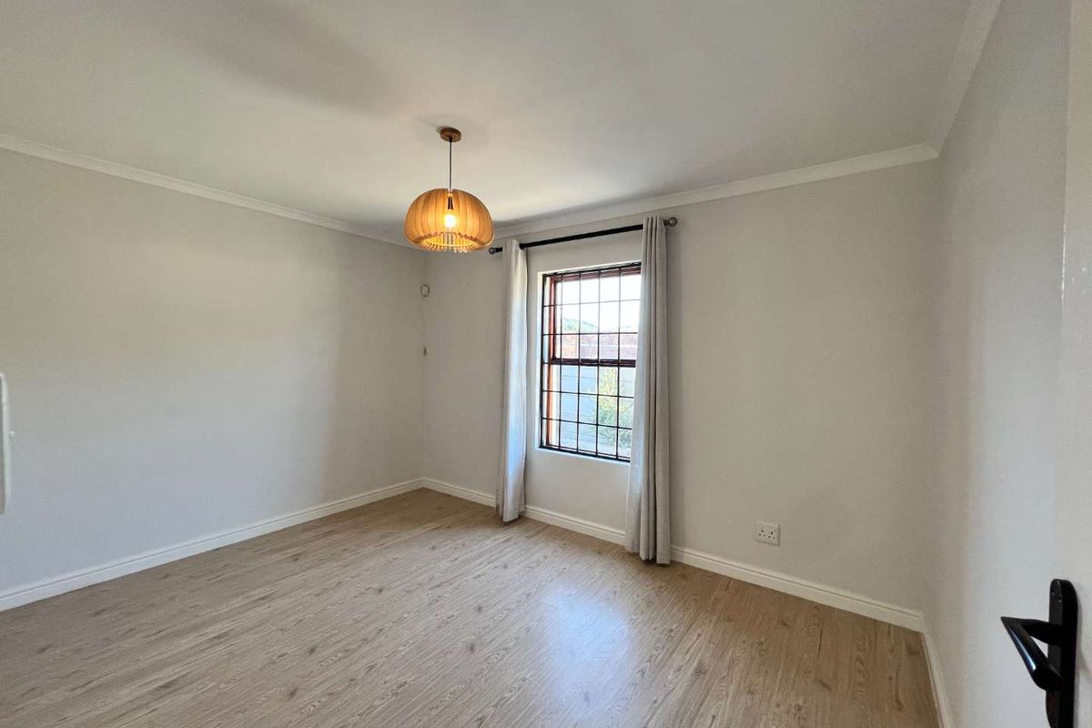 A bright, empty bedroom featuring light-toned laminate flooring, neutral walls, and a window fitted with security bars.