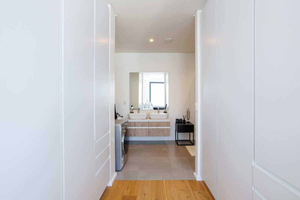 A modern bathroom featuring a double vanity with wood-finish cabinetry, a large mirror, and tiled flooring, viewed from an adjacent hallway.
