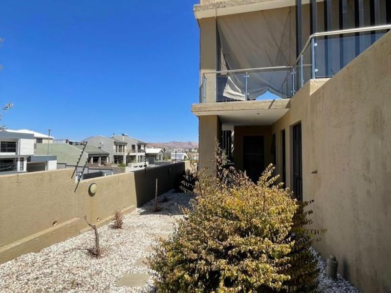 Rear exterior view of the property featuring a gravel garden, retaining wall with electric fencing, and an upper-level balcony with a glass balustrade.
