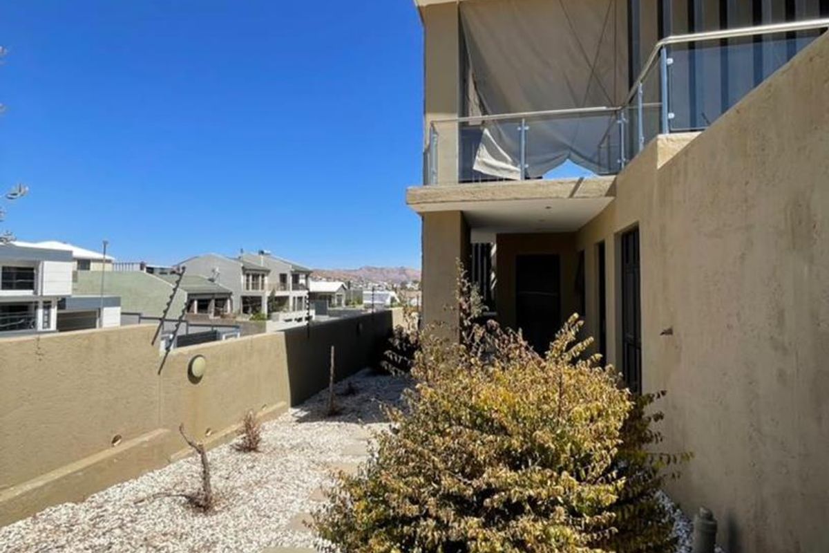 Rear exterior view of the property featuring a gravel garden, retaining wall with electric fencing, and an upper-level balcony with a glass balustrade.