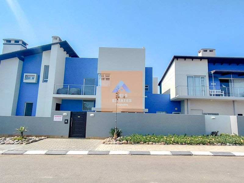 Front elevation of a modern two-story residential property featuring blue and grey facade, balconies, and a paved driveway with a secure boundary wall.
