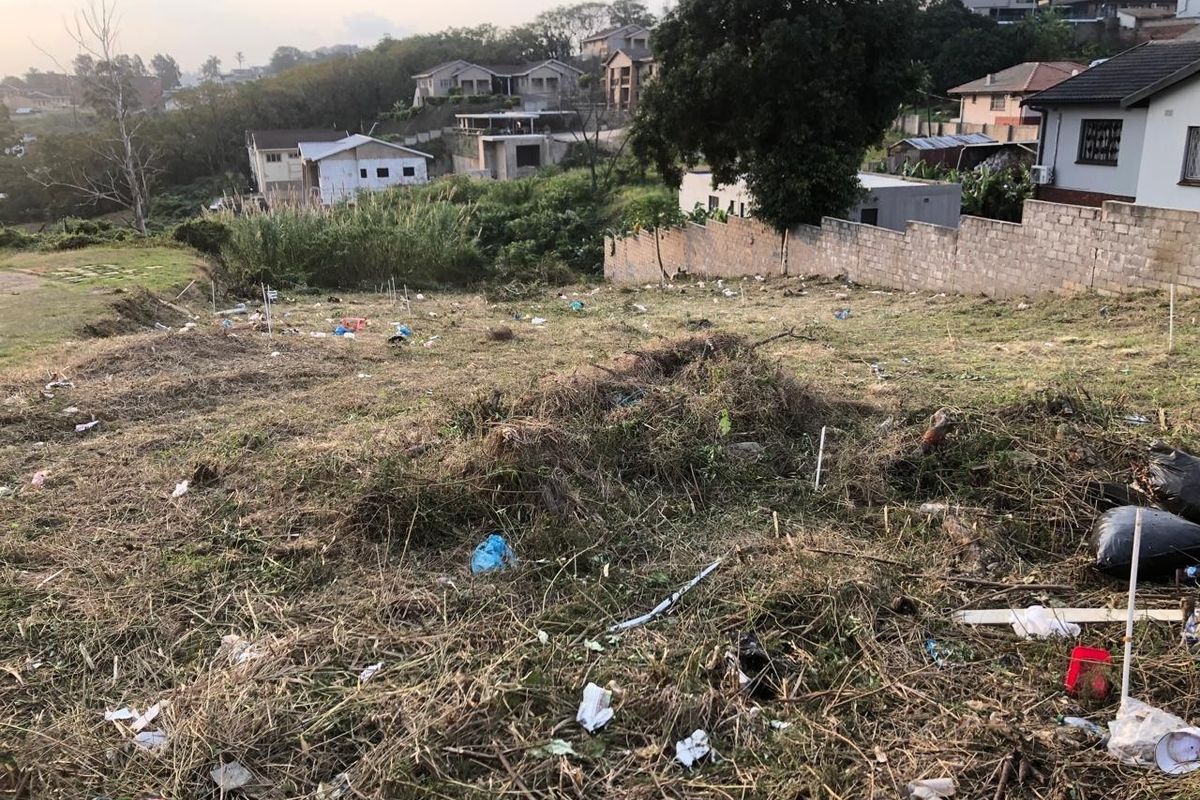 A sloped vacant residential plot featuring a boundary wall and surrounding neighborhood views, currently requiring clearing of vegetation and debris.