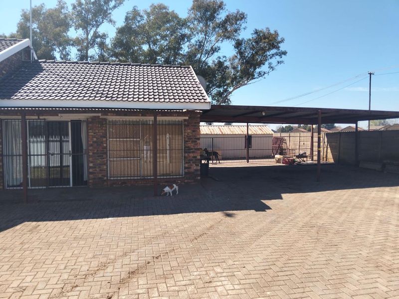 Rear exterior view of the house featuring a brick facade, tiled roof, and a large paved area with an attached carport structure.