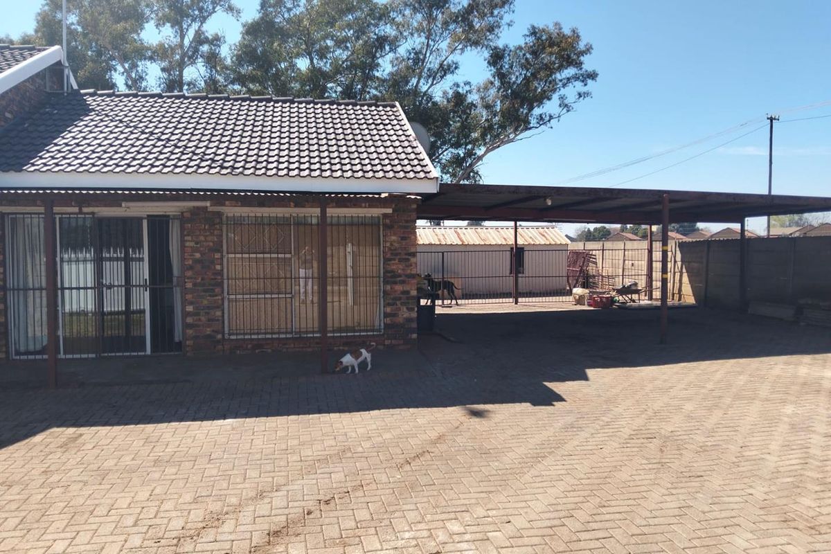 Rear exterior view of the house featuring a brick facade, tiled roof, and a large paved area with an attached carport structure.