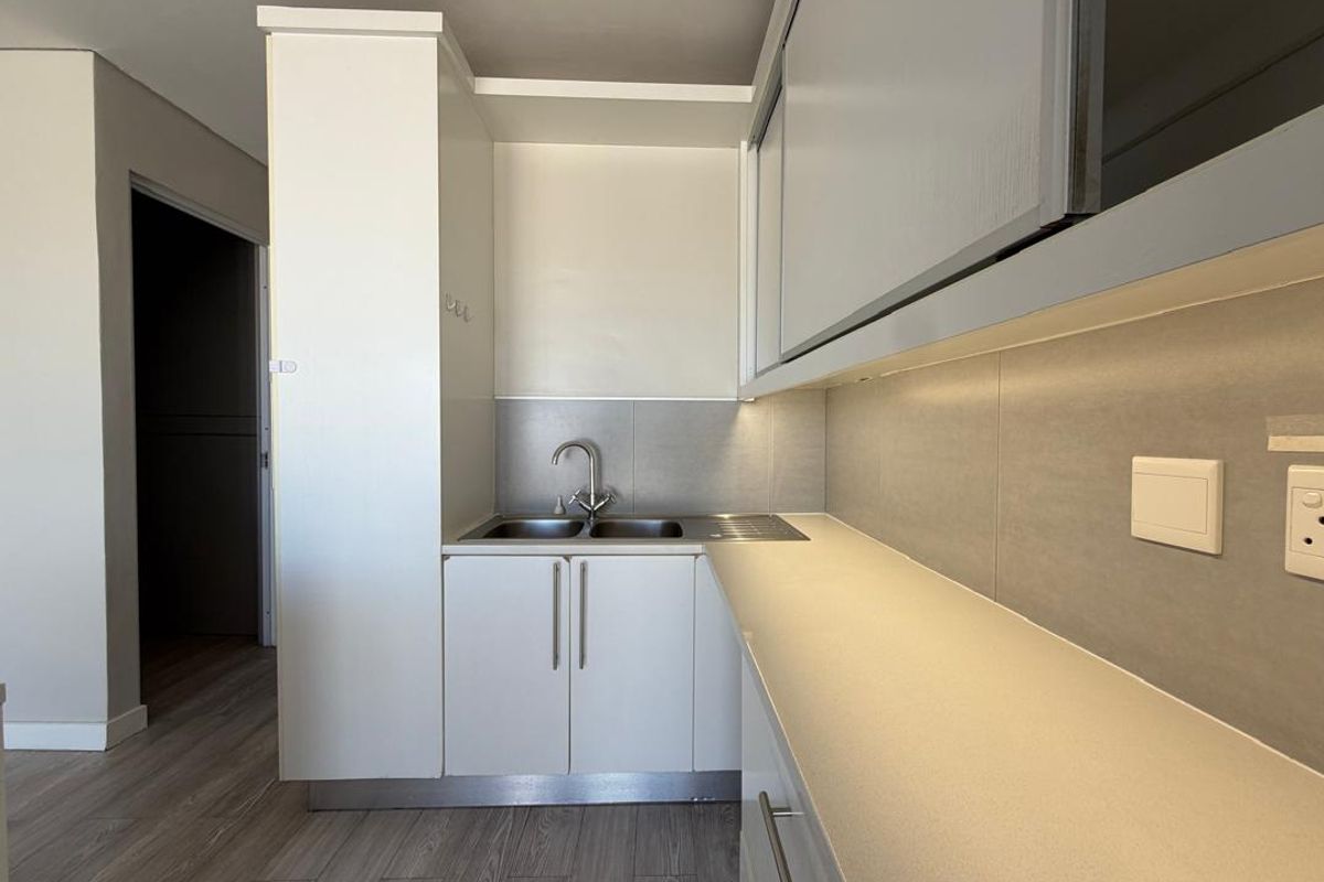 A modern, compact kitchen area featuring sleek white cabinetry, a stainless steel sink, and neutral tiled backsplash with integrated under-cabinet lighting.