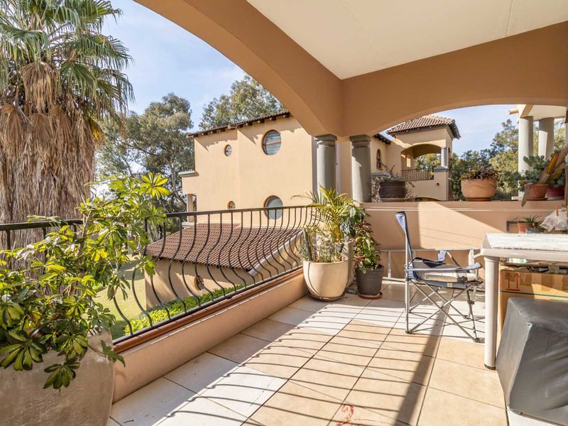 A spacious covered balcony featuring tiled flooring, decorative columns, and a wrought iron balustrade, offering a pleasant outdoor area with garden views.