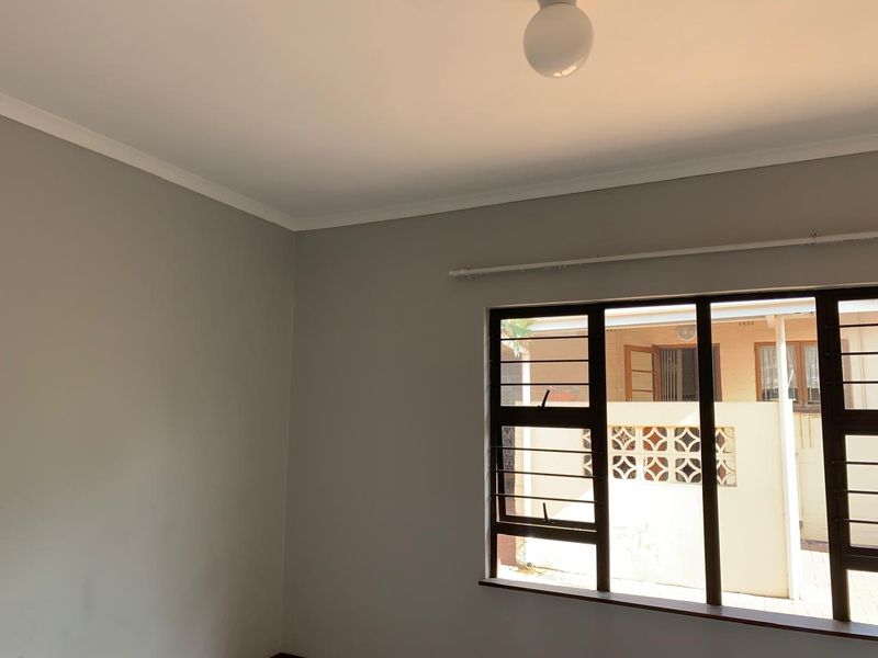 A clean, empty bedroom featuring neutral walls, a metal-framed window with security bars, and a simple ceiling light fixture.