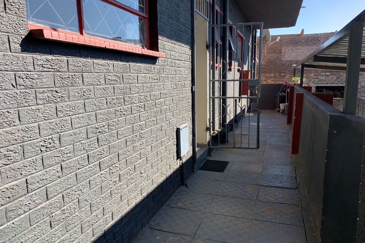 A view of the exterior walkway featuring dark brick walls, metal security gate, and tiled flooring leading to the apartment entrance.