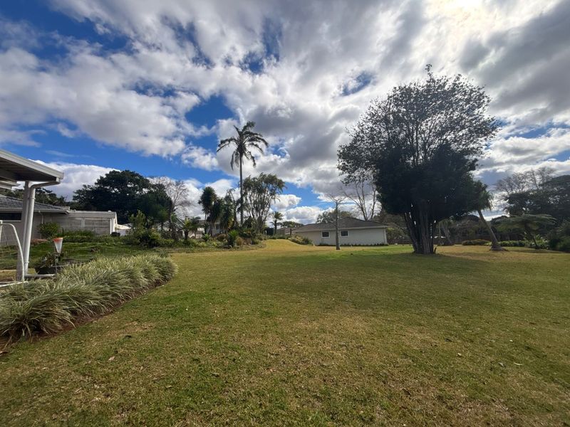 A spacious, well-maintained garden area featuring mature trees, expansive lawn, and a covered patio section attached to the main dwelling.