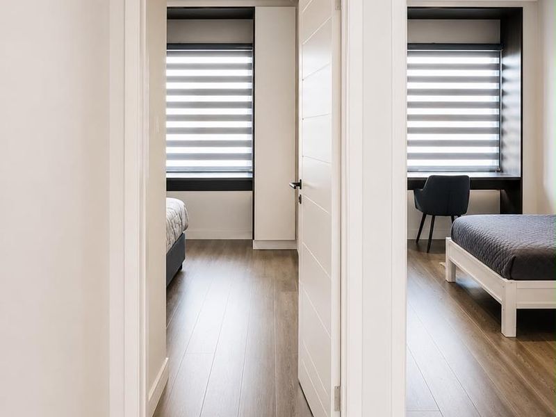 A view from the hallway looking into two modern bedrooms featuring laminate flooring, built-in cabinetry, and integrated desk spaces with roller blinds.