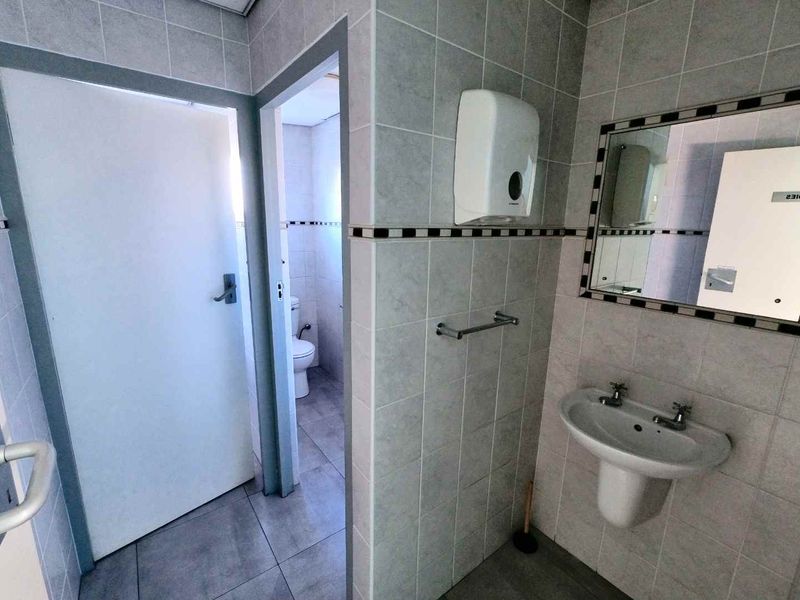 A functional office bathroom featuring tiled walls and flooring, a pedestal washbasin, and a wall-mounted mirror.