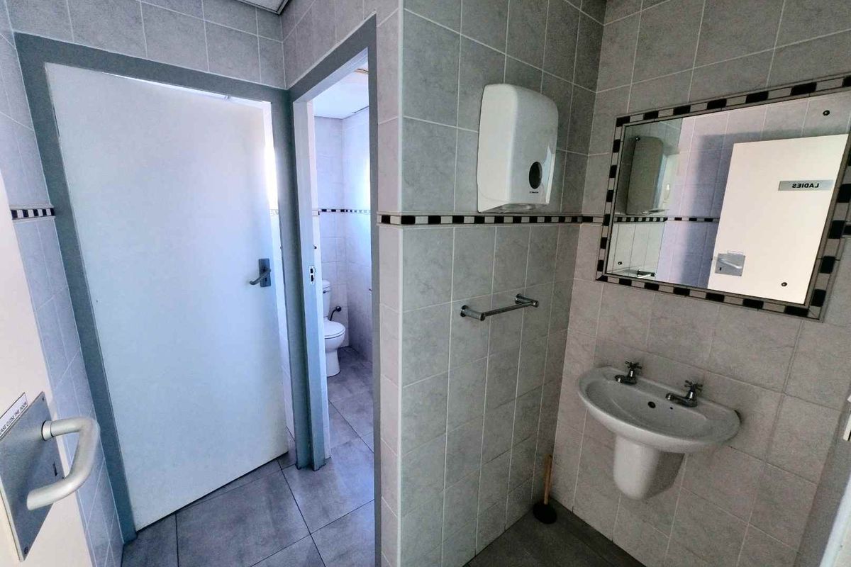 A functional office bathroom featuring tiled walls and flooring, a pedestal washbasin, and a wall-mounted mirror.