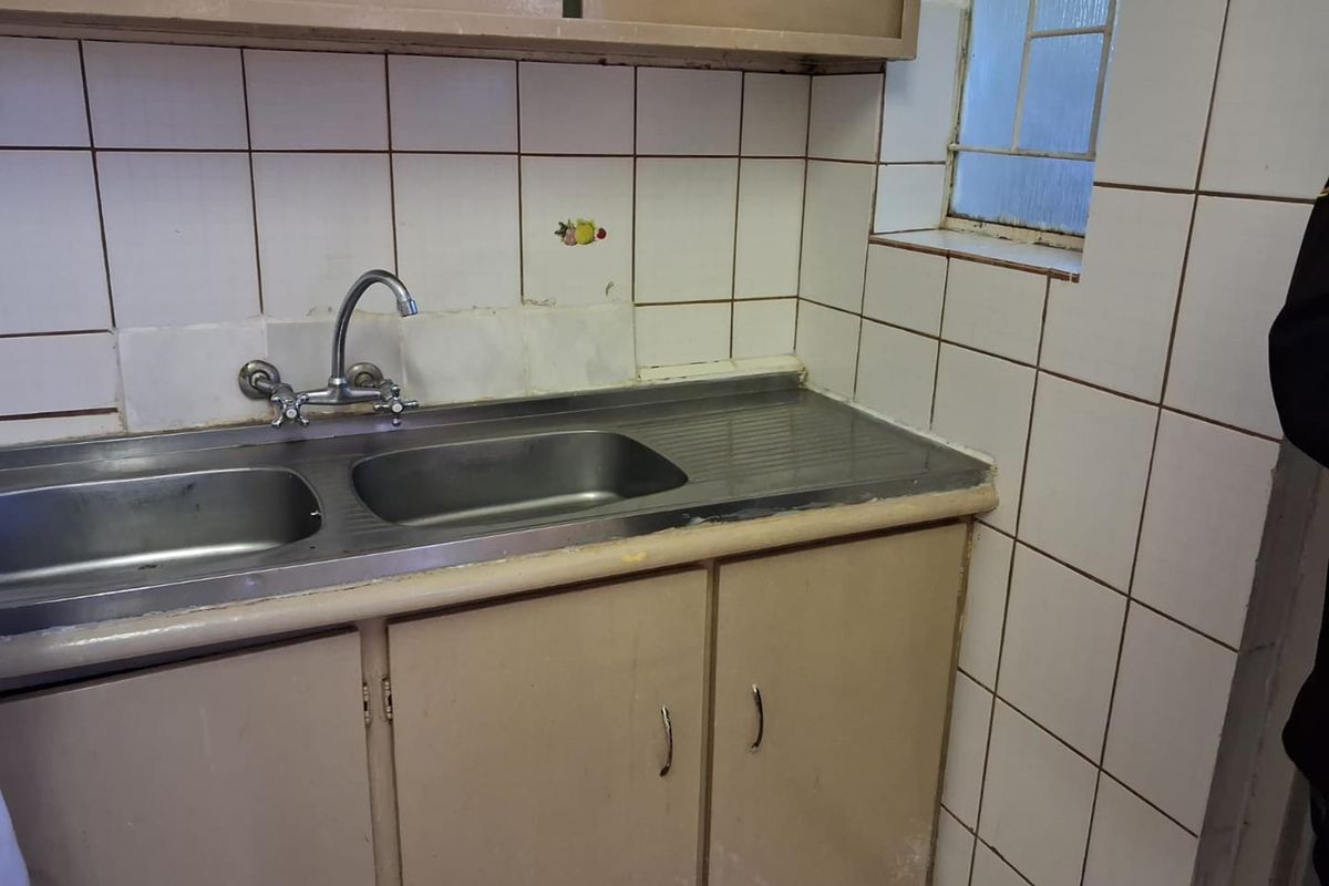 A dated kitchen area featuring a stainless steel double sink, tiled splashback, and original built-in cabinetry in need of modernization.