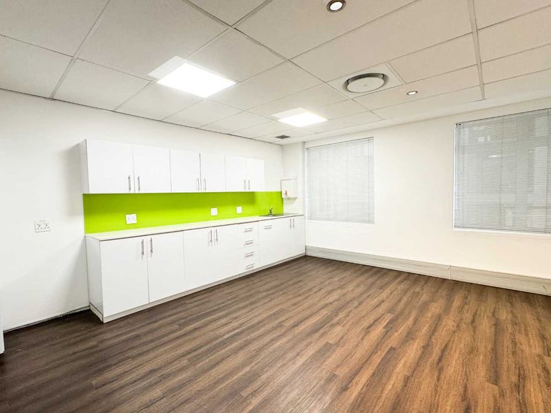 A clean, modern office kitchenette featuring white cabinetry, a vibrant green splashback, wood-look laminate flooring, and recessed ceiling lighting.