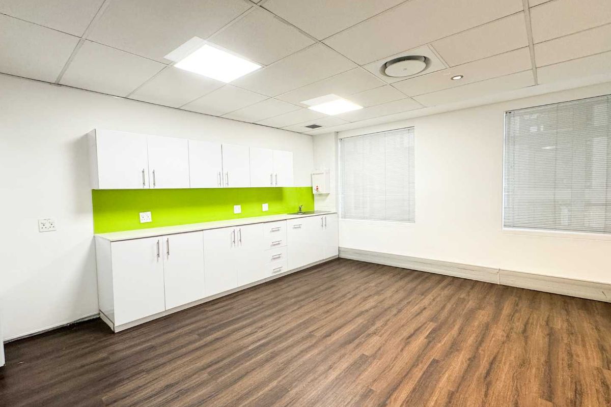 A clean, modern office kitchenette featuring white cabinetry, a vibrant green splashback, wood-look laminate flooring, and recessed ceiling lighting.