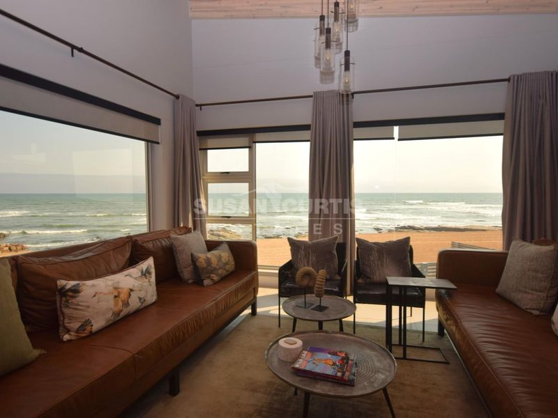 A bright living room featuring high ceilings with exposed timber beams and expansive floor-to-ceiling windows offering panoramic ocean views.
