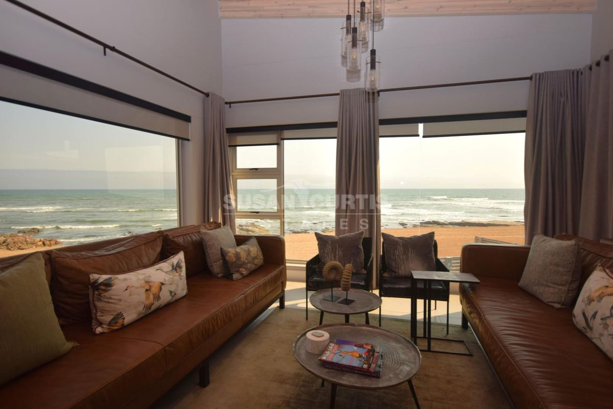 A bright living room featuring high ceilings with exposed timber beams and expansive floor-to-ceiling windows offering panoramic ocean views.