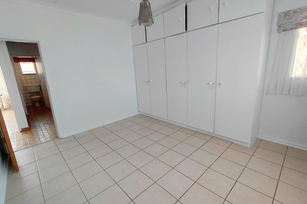 A bright, empty bedroom featuring neutral tiled flooring and extensive white built-in cupboards, with an adjacent view into a tiled bathroom.