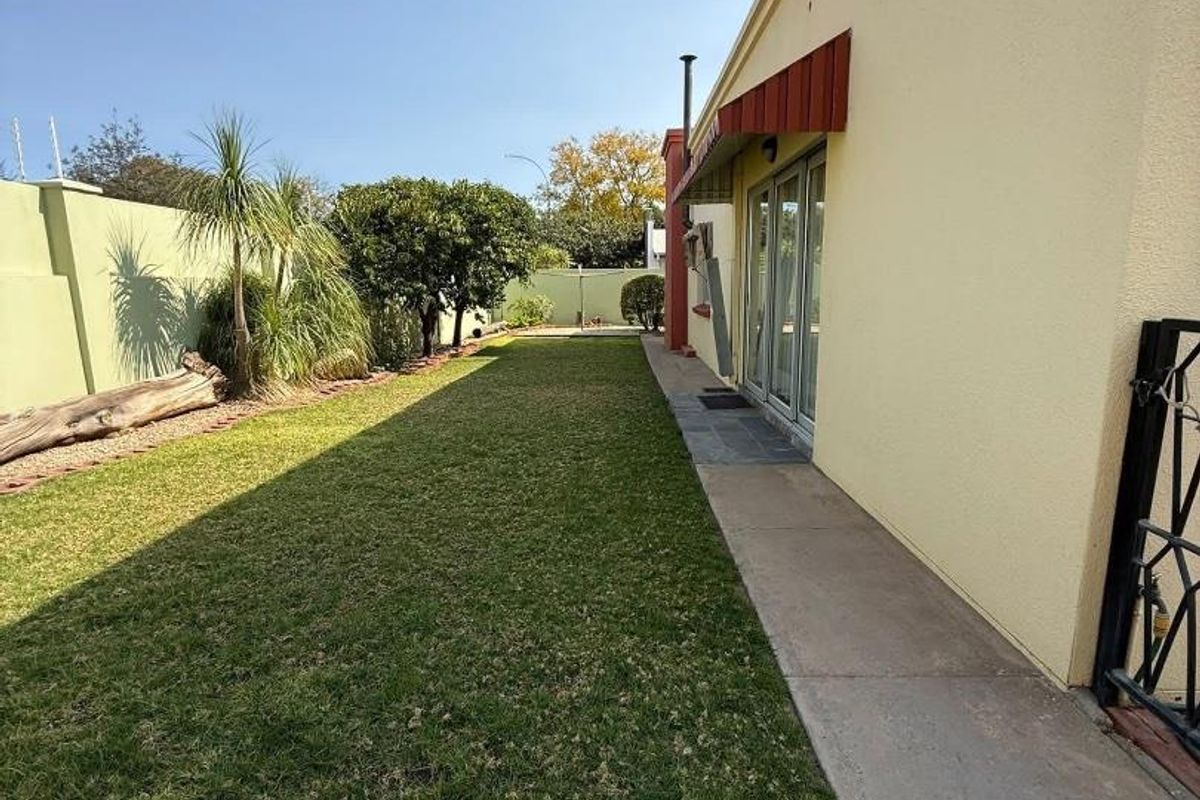 A side garden area featuring a paved walkway, manicured lawn, perimeter wall, and a building exterior with a red awning.